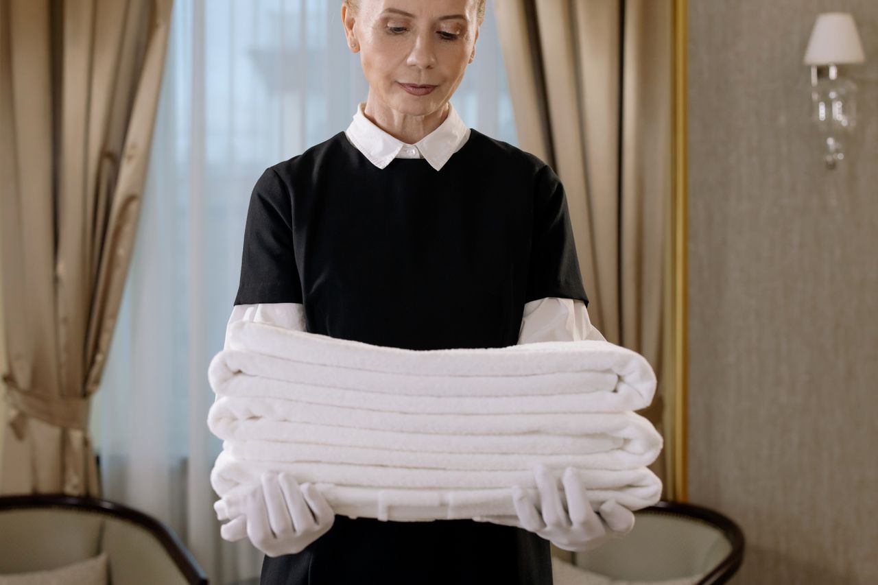 services-01 Professional housekeeper holding a stack of fresh towels in a luxurious hotel room, highlighting elegance and attentive service.